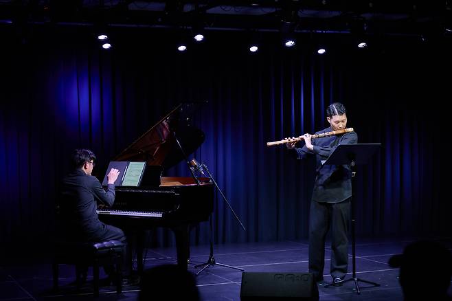 어두운 무대 위에서 한 남성이 피리를 연주하고 있고, 다른 남성은 피아노를 치고 있는 사진이다.