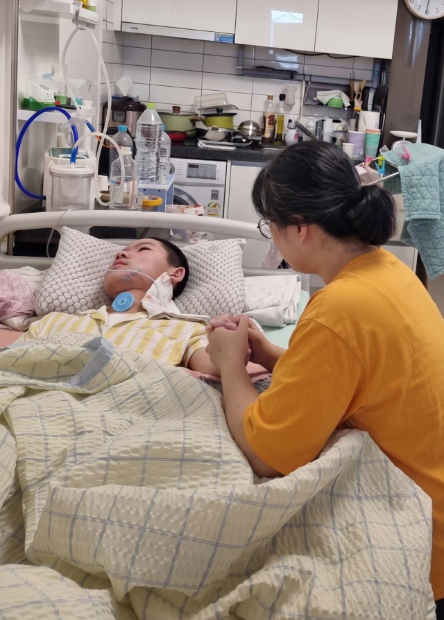 ▲5평 원룸에서 아들 민건이의 곁을 지키며 굳은 손을 꼭 쥐어주는 엄마 박현신 씨. 단 한 순간도 포기하지 않은 엄마의 간절함이 세상에서 가장 따뜻한 온기가 되어 아이에게 스며들고 있다. ⓒ프레시안